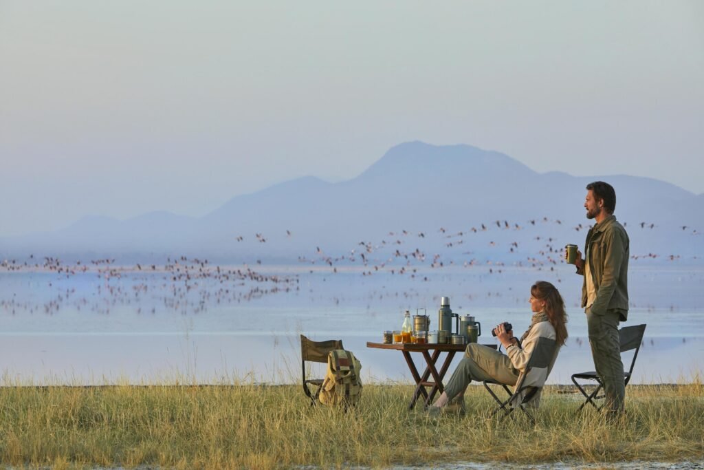 two people sit at a table by a lake drinks and cameras on the table while a man stands holding a cup mountains in the distance