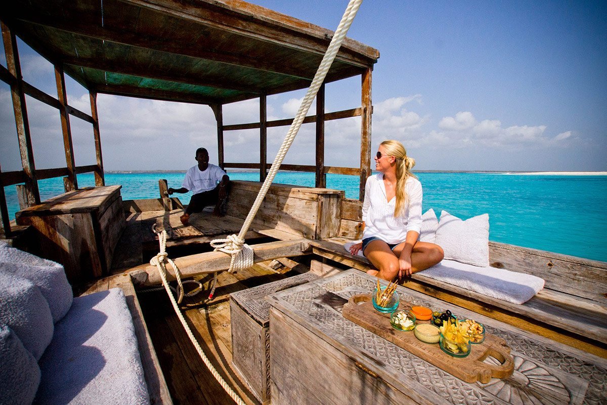 Dhow-Cruise-Camping-Tours-Sinda-Island-at-Kigambonijpg - EXCELLENCE AFRICA ADVENTURES - EXCELLENCE AFRICA ADVENTURES woman in a white shirt sits on a sunlit wooden boat deck with turquoise sea in the background a man nearby in the shadows made by the shelter