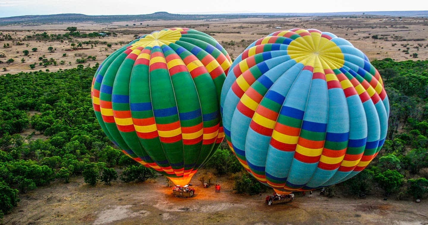 balloon-flight-masai-mara-national-reserve-3jpg - EXCELLENCE AFRICA ADVENTURES - EXCELLENCE AFRICA ADVENTURES two large colorful hot air balloons on the ground baskets attached preparing for takeoff in a dry open landscape with sparse trees