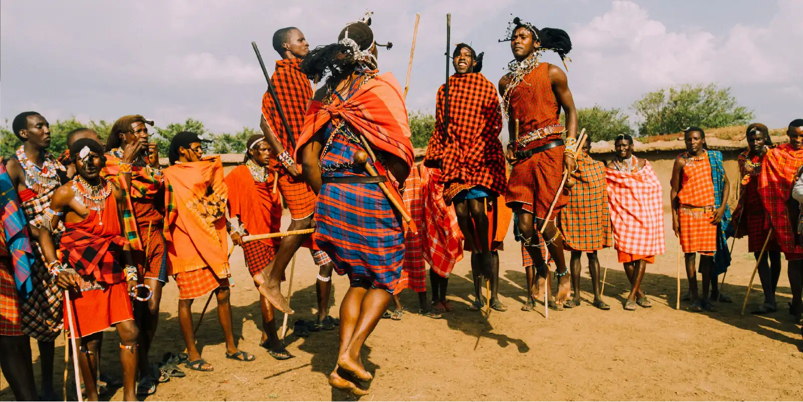 cultural-safari-herowebp - EXCELLENCE AFRICA ADVENTURES - EXCELLENCE AFRICA ADVENTURES maasai people in red shukas perform a jumping traditional dance with sticks in a dusty village setting