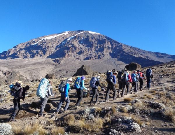 Group of hikers marching along a rocky trail toward a rugged mountain with patches of snow under a clear blue sky.