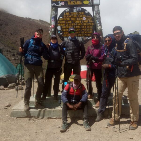 group of hikers posing by the kilimanjaro national park sign in tanzania with tents and rocky terrain behind them