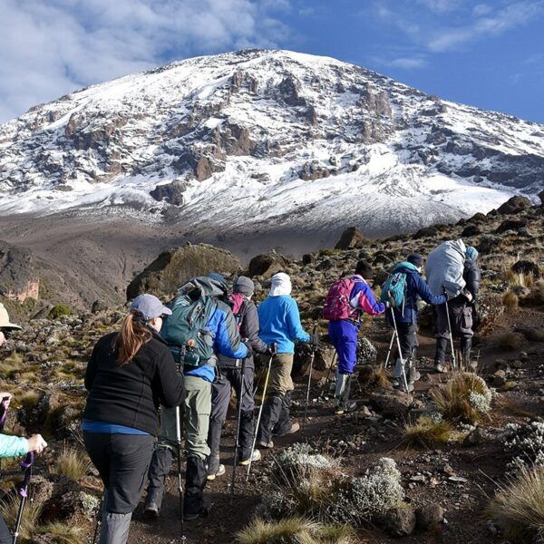 Group of hikers with trekking poles ascending a rocky trail toward a snow-capped mountain under a blue sky.